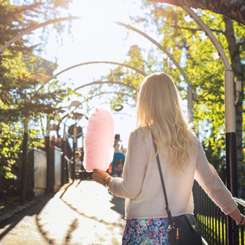 Blonde woman holding cotton candy walks through a sunlit archway in a park.