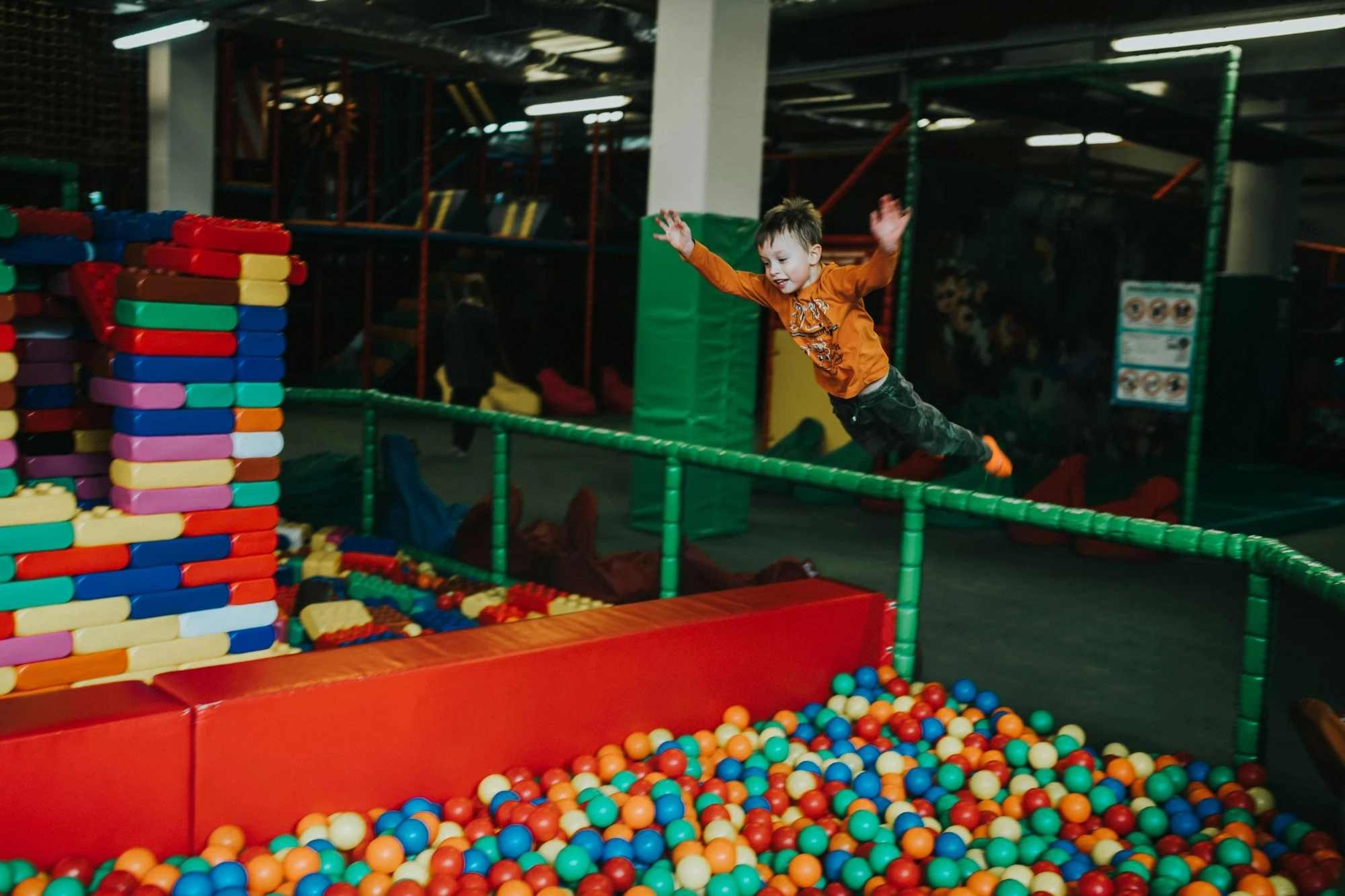 A child joyfully jumps into a colorful ball pit at an indoor playground. Wearing an orange shirt, he looks excited and carefree against the vibrant backdrop.