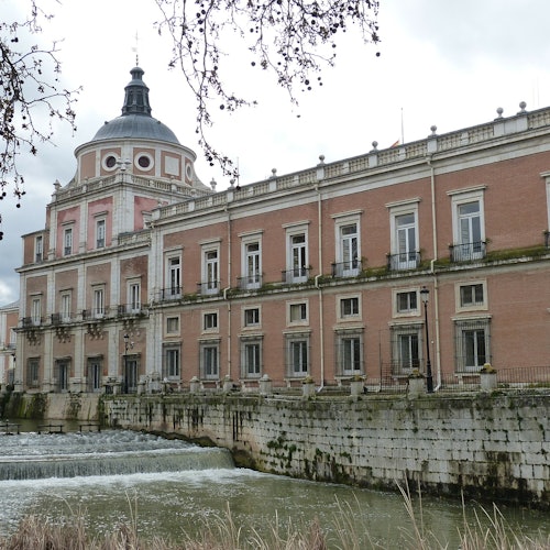 Tarihi bina, kubbeli, pembe tuğla cepheli ve çok sayıda penceresi olan, bulutlu bir gökyüzü altında akan bir nehrin yanında yer almaktadır.