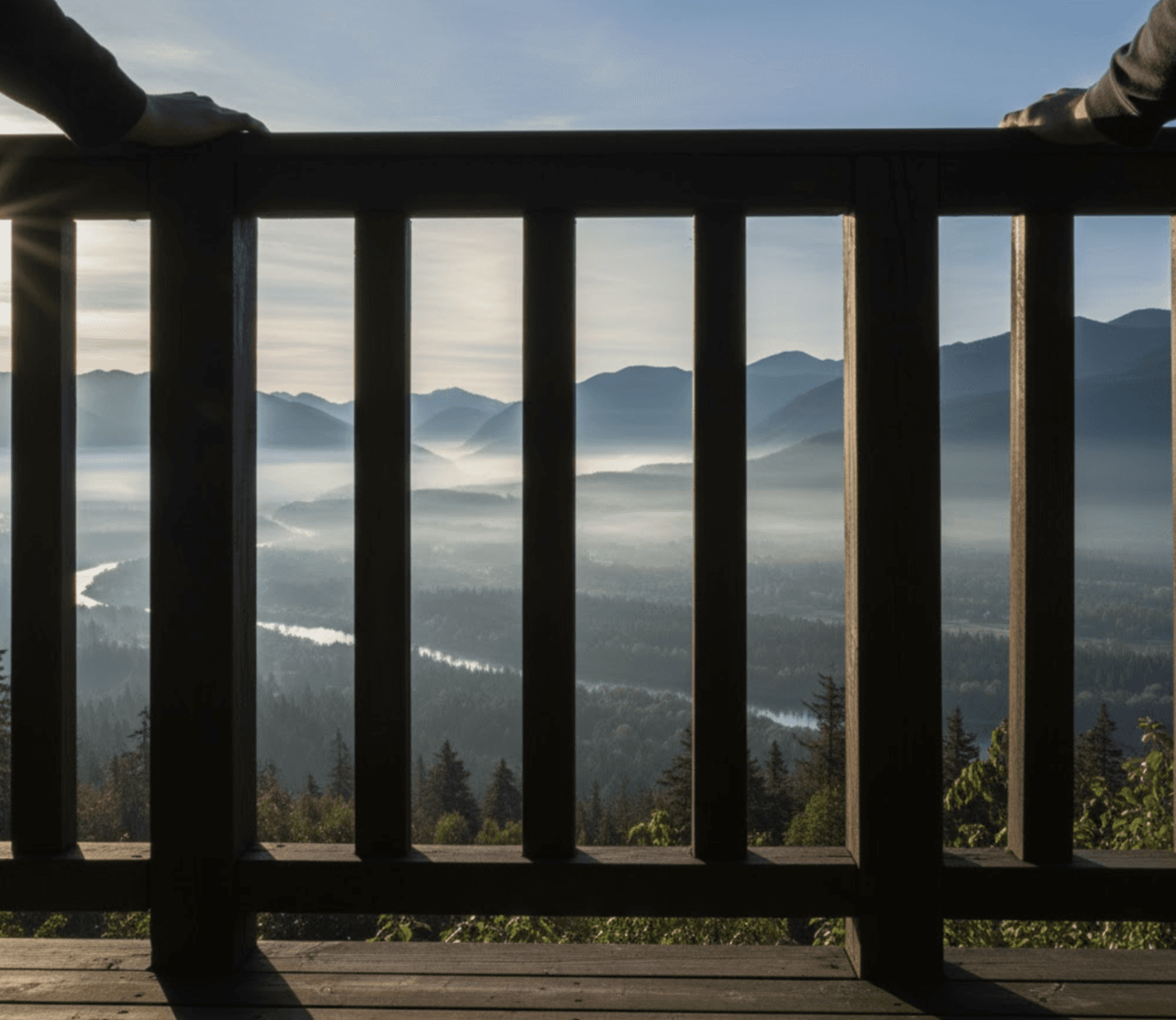 A first-person perspective shot from a small deck overlooking a breathtaking, misty Fraser Valley mountain and river landscape at dawn.