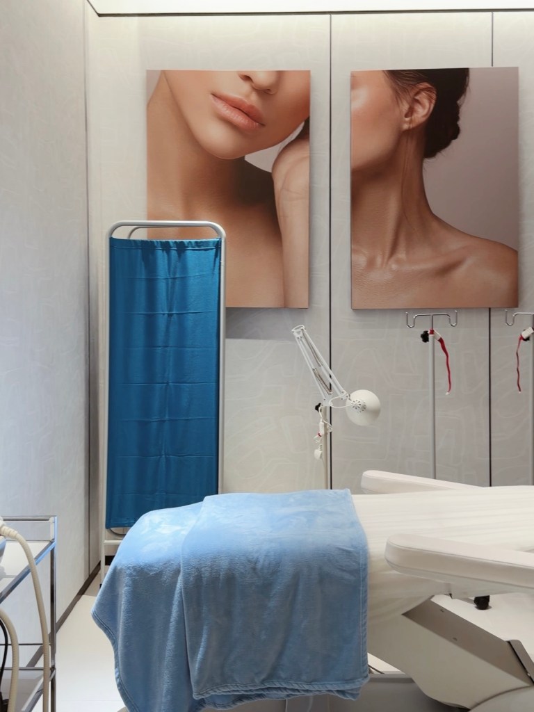A soothing aesthetic clinic room featuring minimalist decor, with large portraits of skin close-ups on the walls, a light blue blanket on a treatment bed, and a medical supply cart.