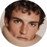 A person wearing a large, fluffy beige scarf wrapped around the neck, accompanied by a brown textured sweater. The expression appears calm and pensive.