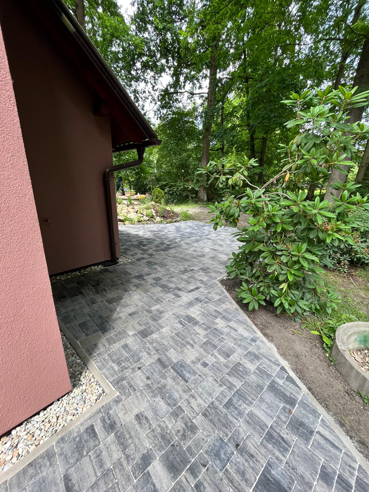 Dlážděná cesta k domu se stěnou v hnědé barvě, s rostlinami po stranách. Venkovní prostředí.