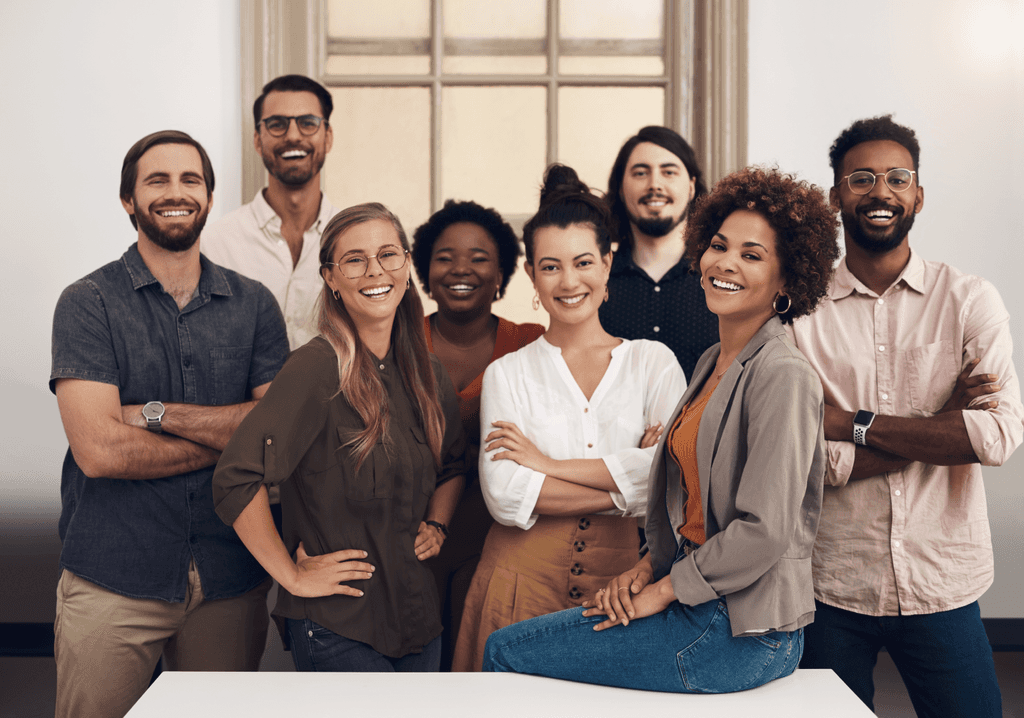 Grupo diverso de pessoas em pé, sorrindo e posando para a foto em frente a uma grande janela de escritório. A imagem transmite um ambiente acolhedor e colaborativo, refletindo a alegria e a união do grupo.