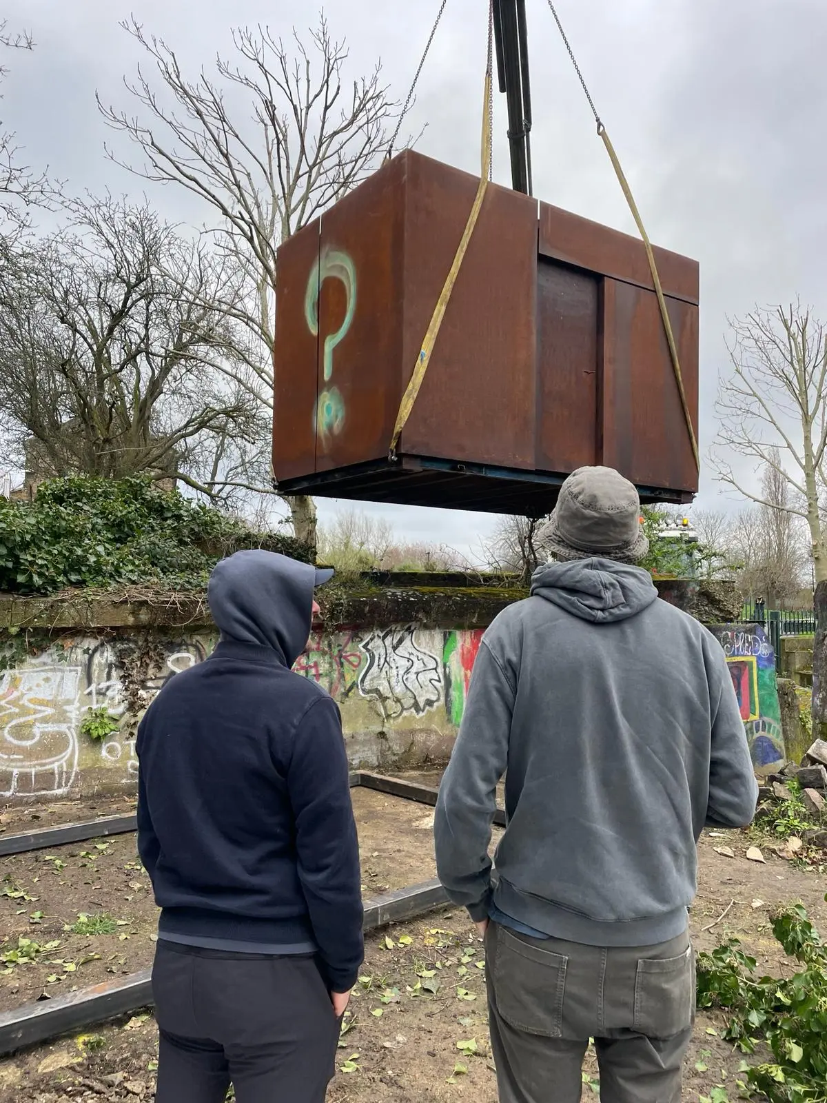 Two people in hooded sweatshirts watch a large brown box suspended from a crane against a cloudy sky.