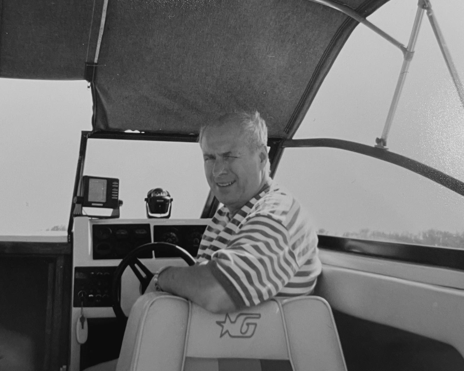 Black-and-white photo of Normy seated at the helm of a boat, smiling as he prepares to retire from his career in home textiles entrepreneurship.