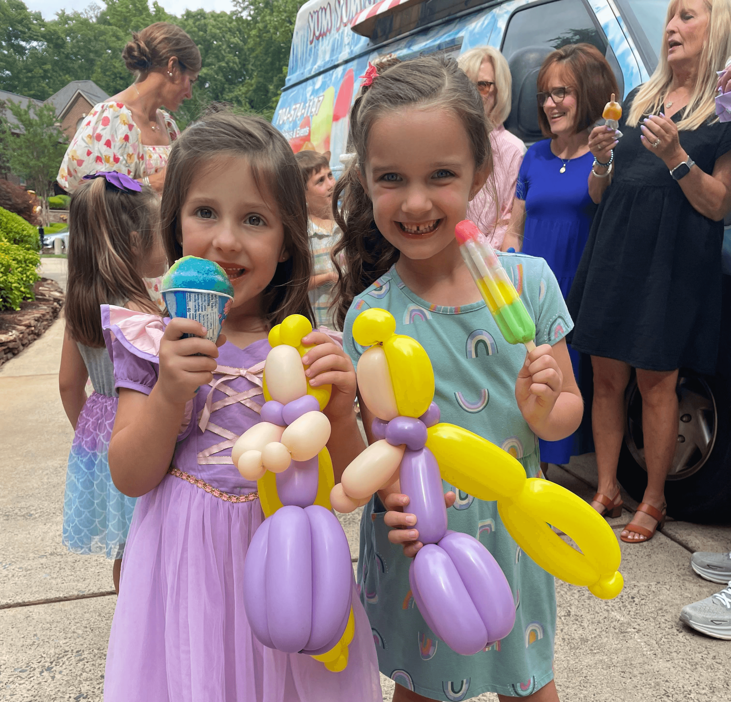 Two young girls smiling and holding balloon sculptures and colorful frozen treats at an outdoor gathering, with a decorated ice cream truck and group of people in the background
