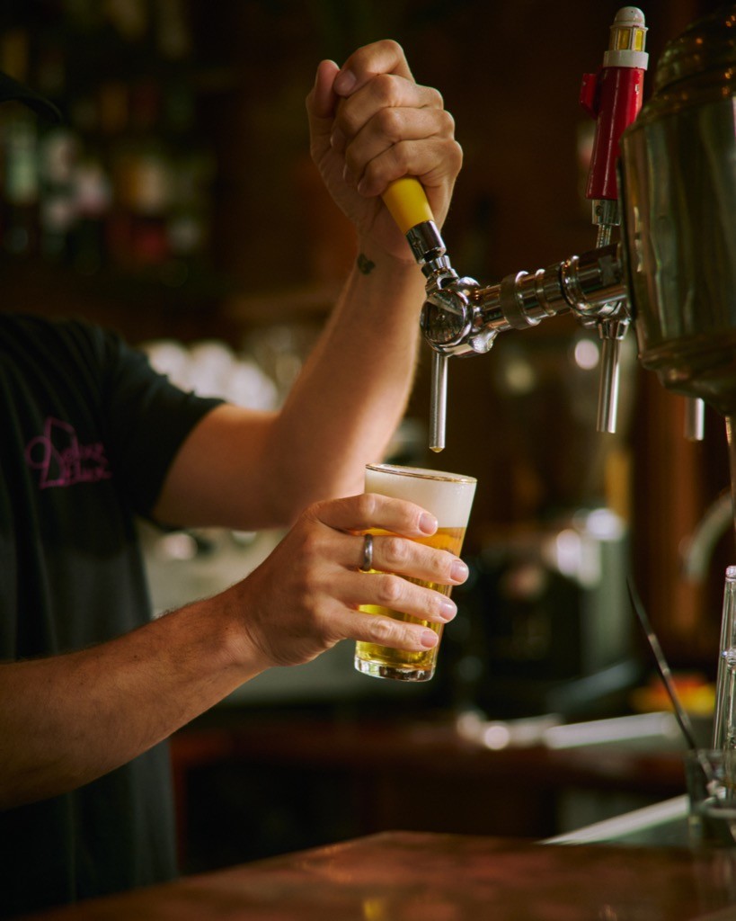 Person pouring a beer from a tap into a glass at a bar, with a dimly lit background and visible bar equipment.