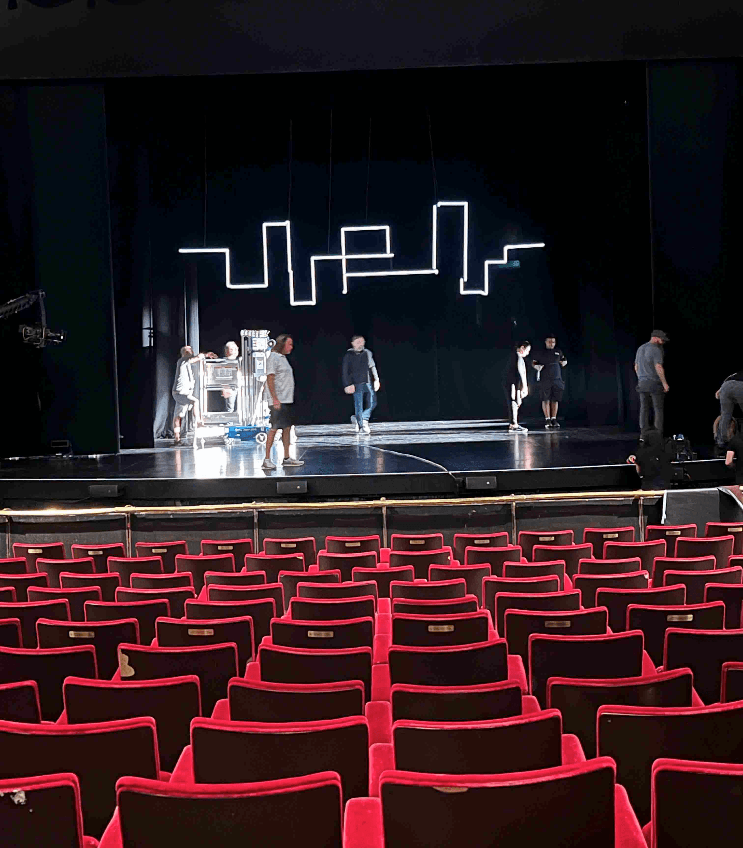 A theater stage set up with performers, viewed from the audience seats with red carpeting.