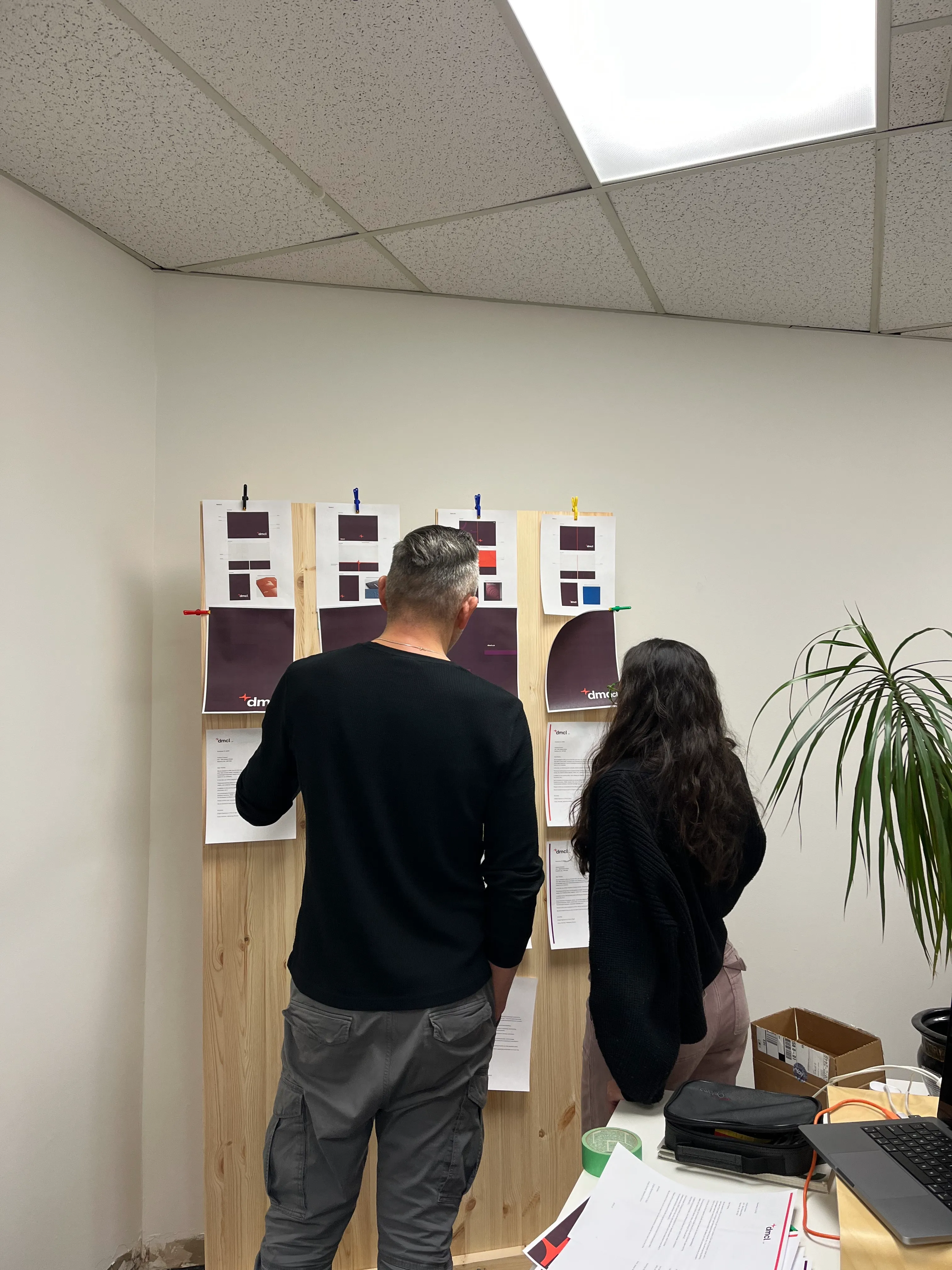Jason Schultz and Alejandra Silva Rojas looking at print outs of concepts for DMCL's brand identity design. They are hangining up on wooden stands in an office.