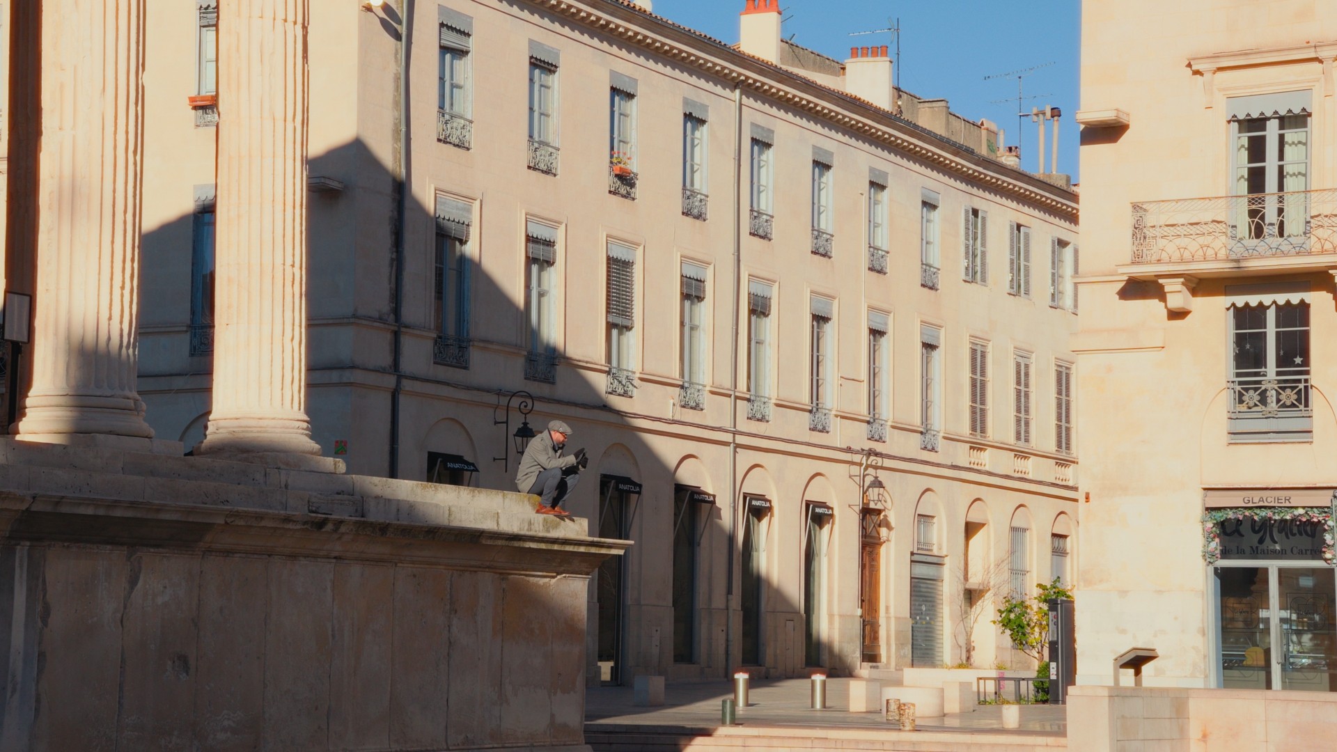 Un homme assis sur un muret en pierre de la Maison Carrée, lisant dans la lumière du matin à Nîmes.