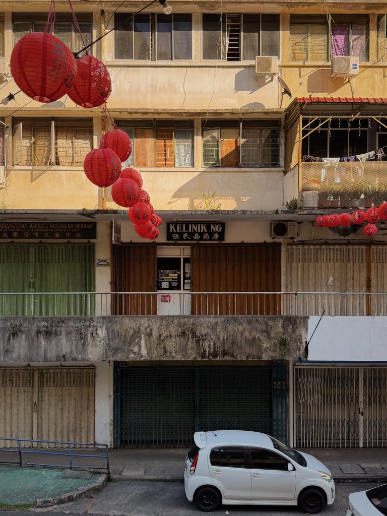 Street view of a building with red lanterns hanging outside, featuring various windows with different shades of colors. A small white car is parked in front of the building.