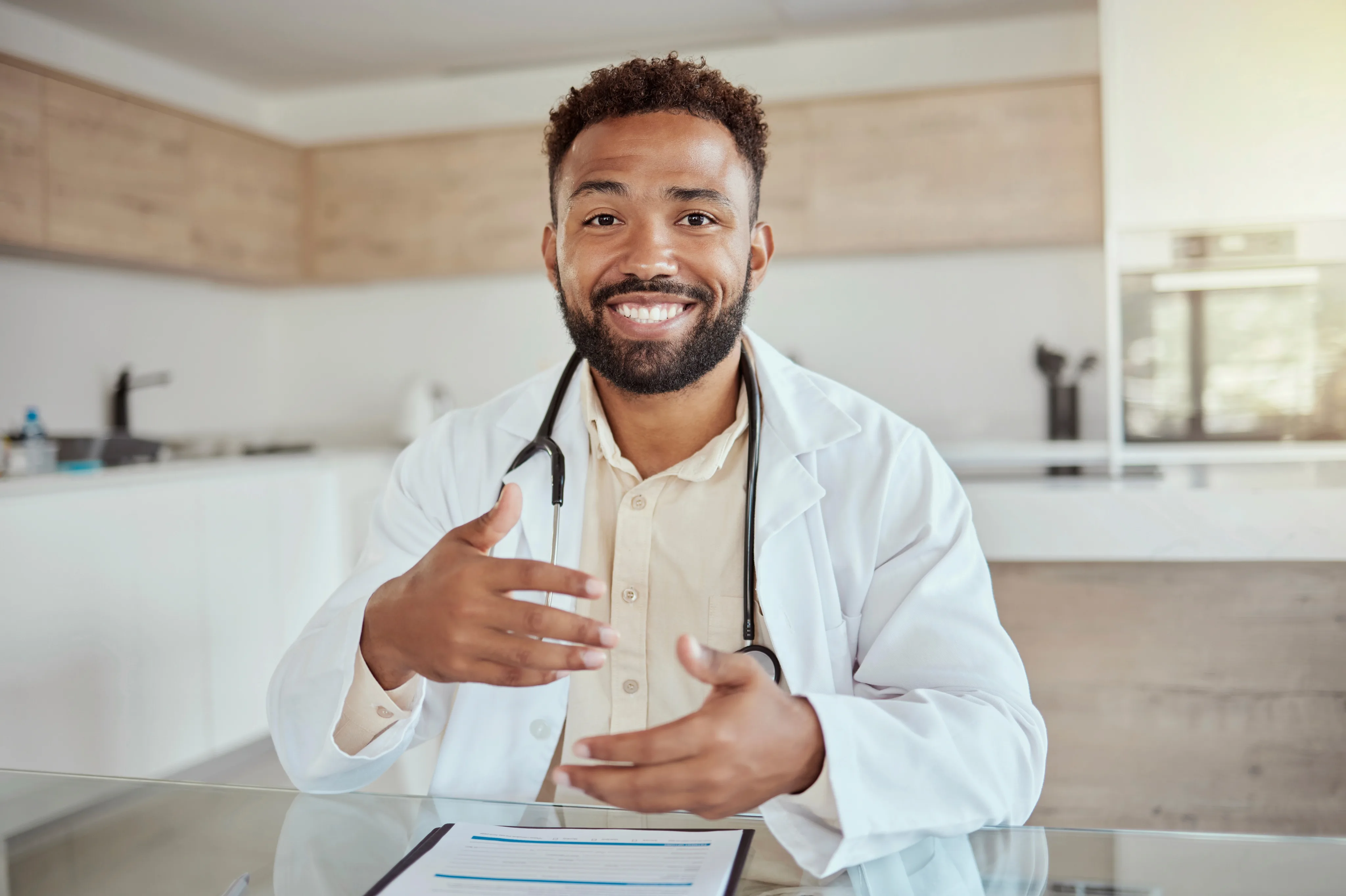 A smiling male doctor in a white lab coat and stethoscope gestures while speaking at a medical clinic desk.