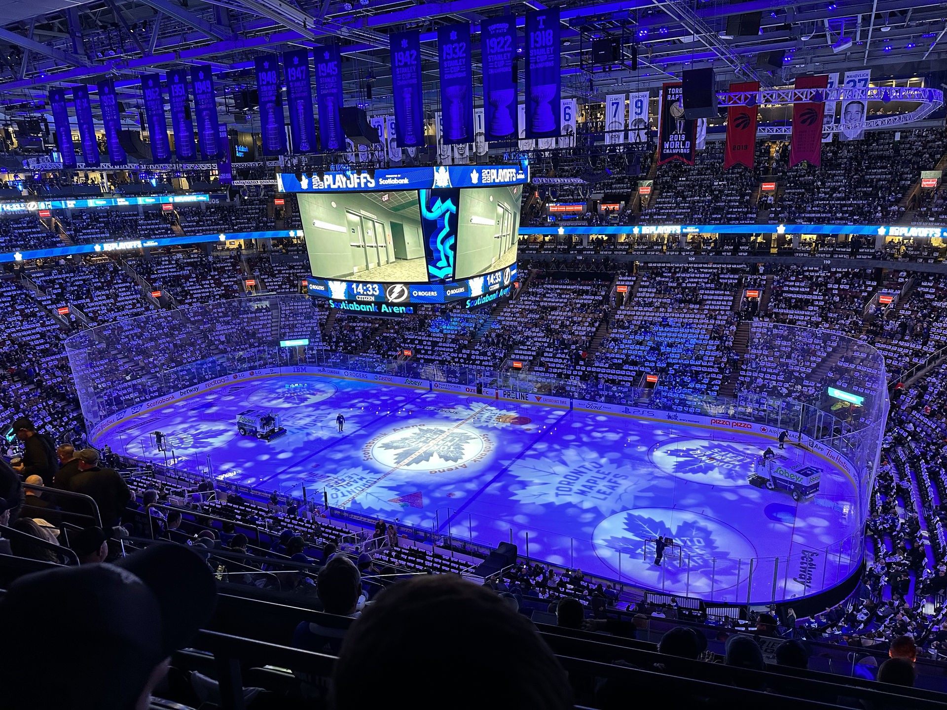 Wide view of a packed hockey arena illuminated in blue lighting, with the Toronto Maple Leafs logo projected onto the ice and playoff graphics on the scoreboard. The crowd is seated and waiting as Zambonis resurface the rink before the game begins.