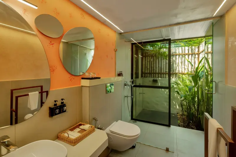 Junior Suite bathroom at Vedic Village Kodungallur with rounded mirrors, indoor vanity and glass-walled outdoor shower opening to a bamboo garden