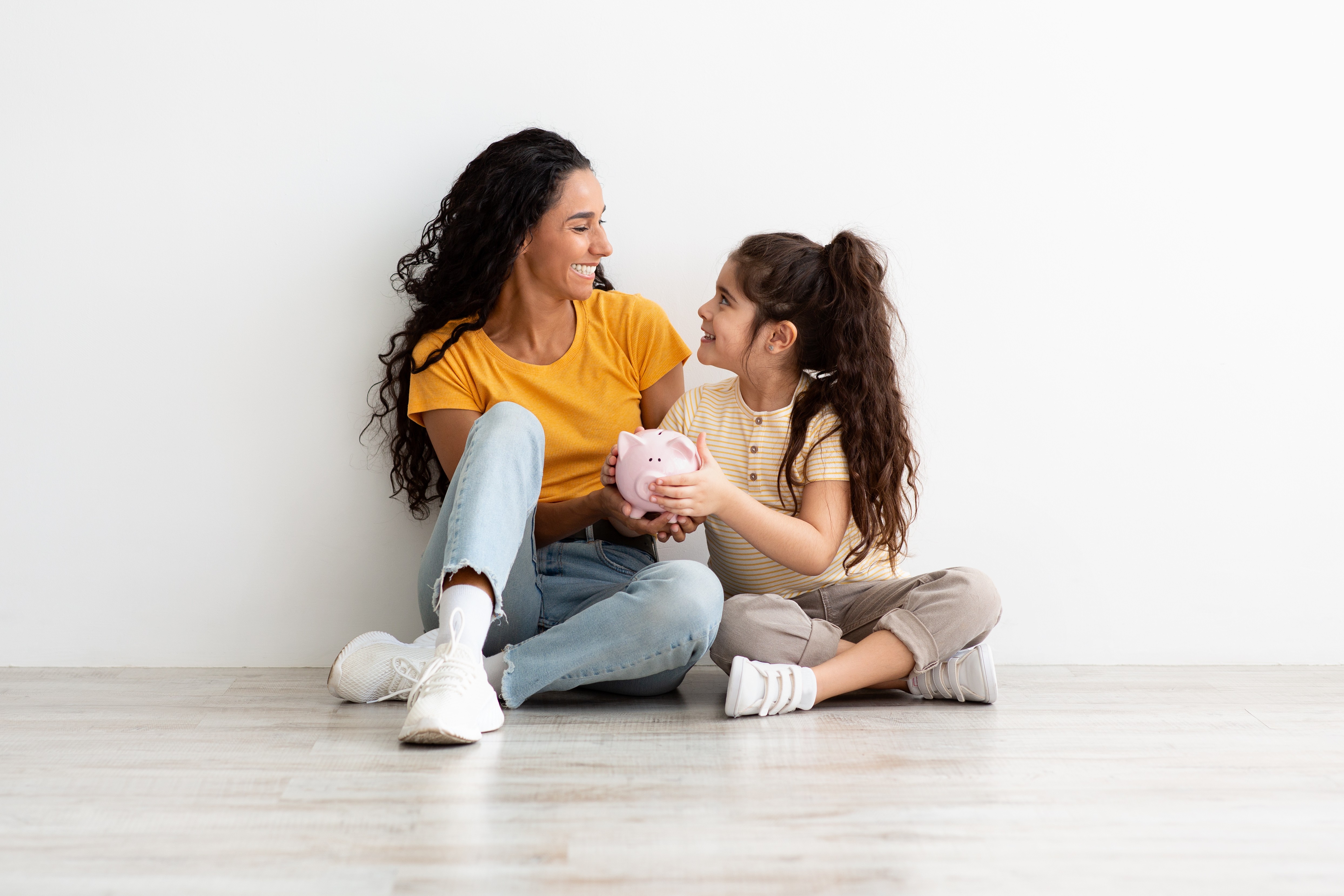 Twee jonge vrouwen zitten op de vloer en delen een vrolijk moment terwijl ze elkaar aankijken en glimlachen tegen een eenvoudige witte muur.