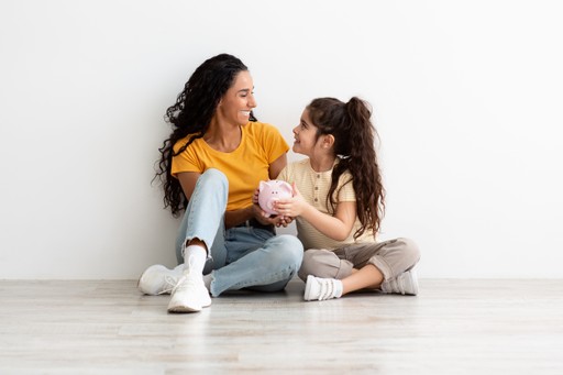 Two young women sit on the floor, sharing a joyful moment and smiling at each other against a plain white wall.