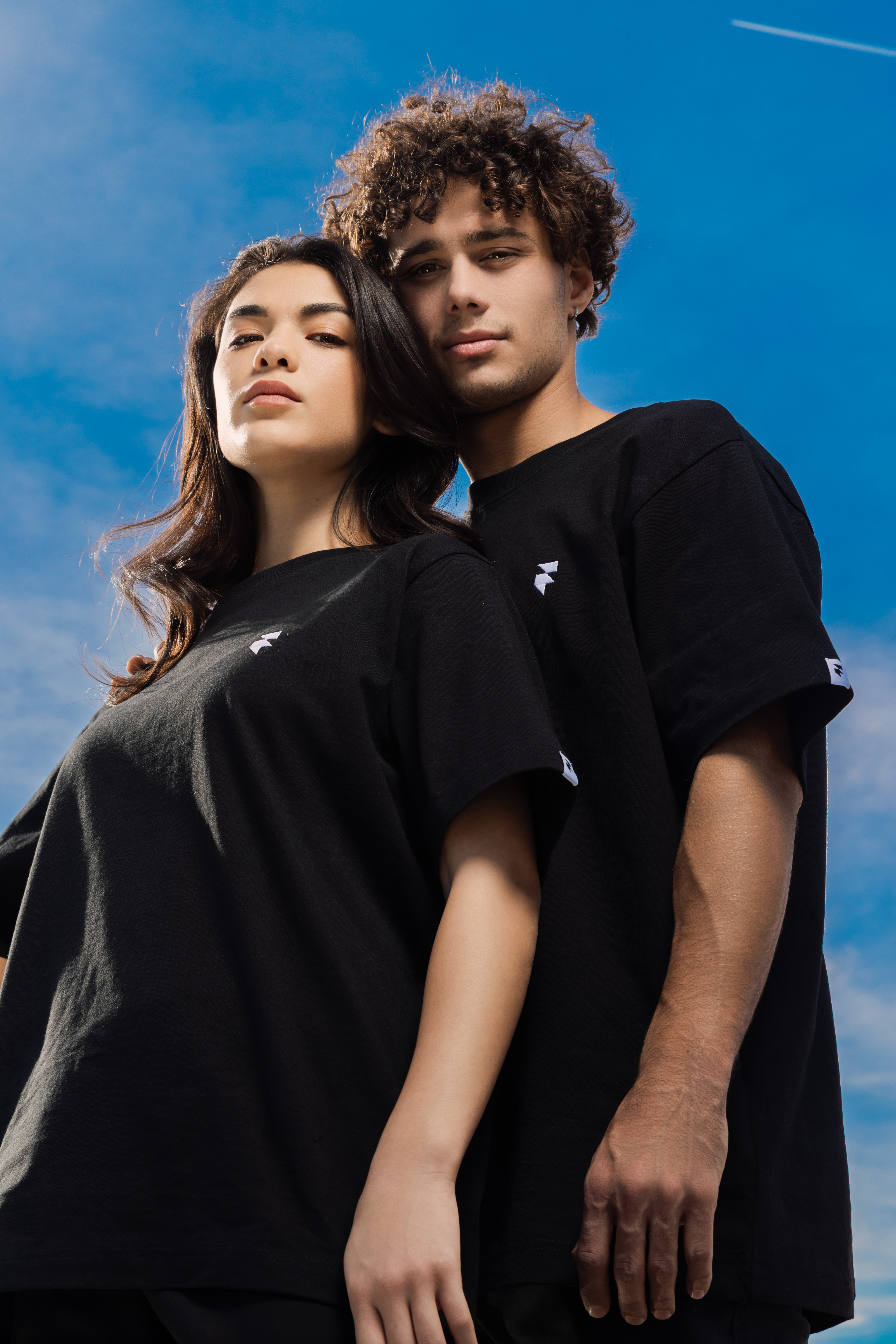 Two individuals wearing black t-shirts stand closely together against a clear blue sky with a contrail in the background.