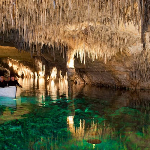People in a small boat glide through a cave with illuminated stalactites and clear, greenish water.