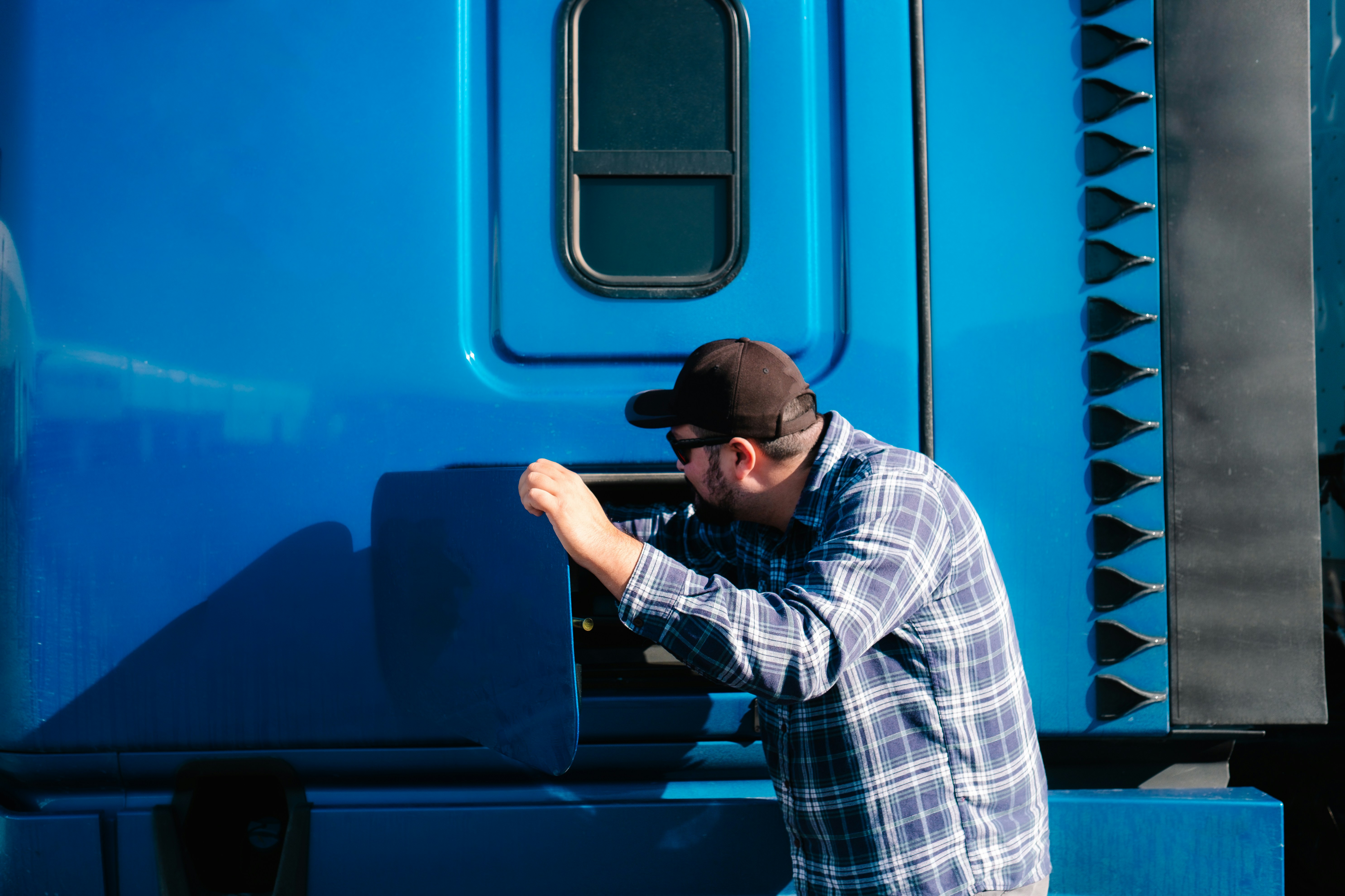 Here's alt text for both images: Image 1 (Truck Driver Checking Truck for Maintenance): "Truck driver wearing a plaid shirt, baseball cap, and sunglasses performing a maintenance inspection on a bright blue semi-truck. He is leaning into an open compartment on the side of the cab, checking mechanical components. Photo by Wesley Tingey on Unsplash.