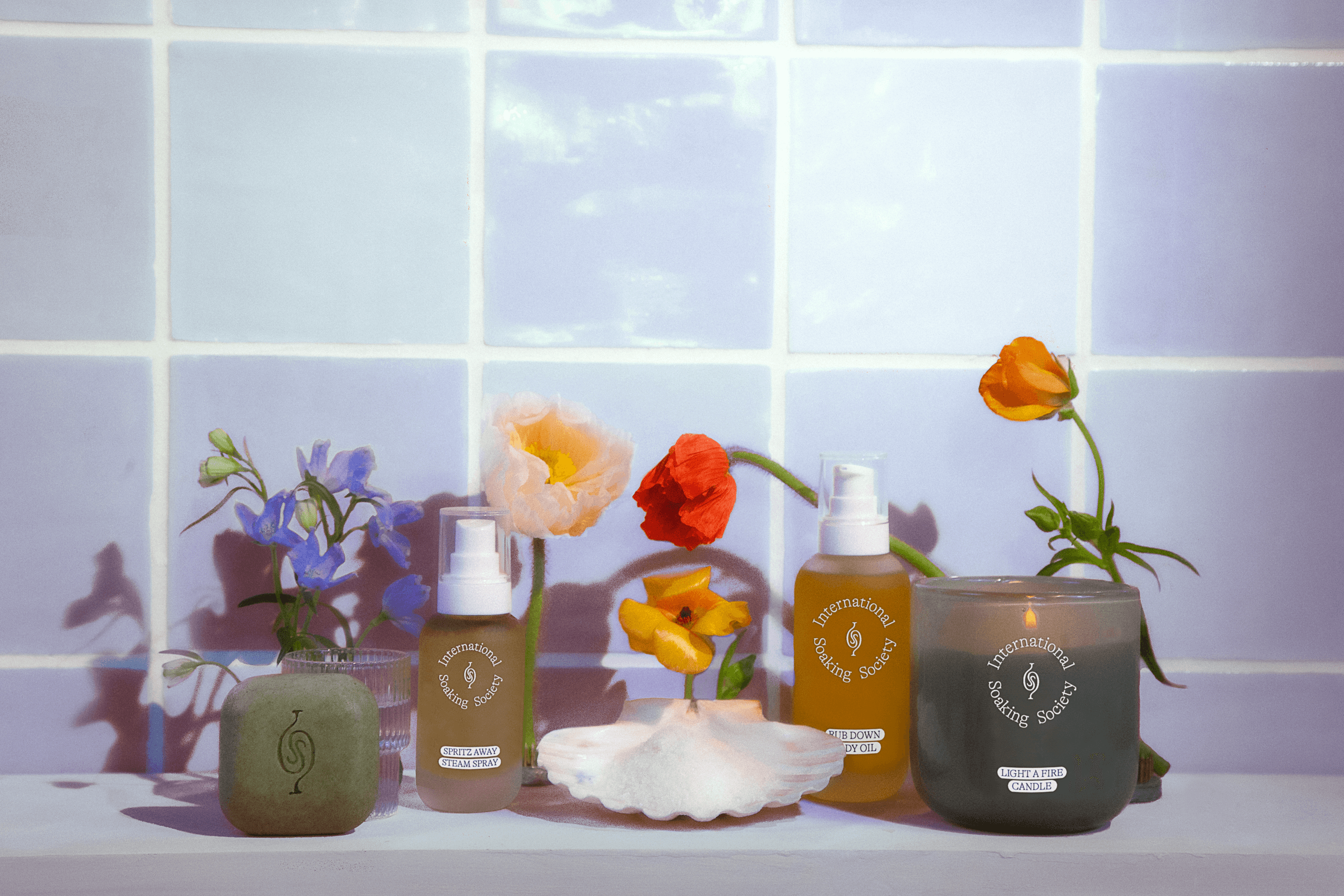 The image shows a collection of six small bottles and two candles arranged on a white surface in front of a light blue tiled wall. The bottles are labeled “Sustainably Sourced Hand Sanitizer” and “Sustainably Sourced Hand Soap”, suggesting they are hand sanitizer and soap. The bottles are various shades of brown and amber. Two of the candles are in a dark green/olive jar and two are in a clear jar; one of the clear jars contains a small arrangement of blue flowers. The wall behind them is covered in square tiles.