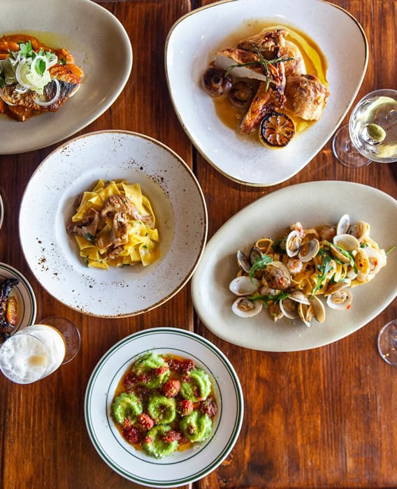 Overhead view of four elegant Italian dishes on a wooden table, including pasta, seafood, and a green appetizer, accompanied by a drink.