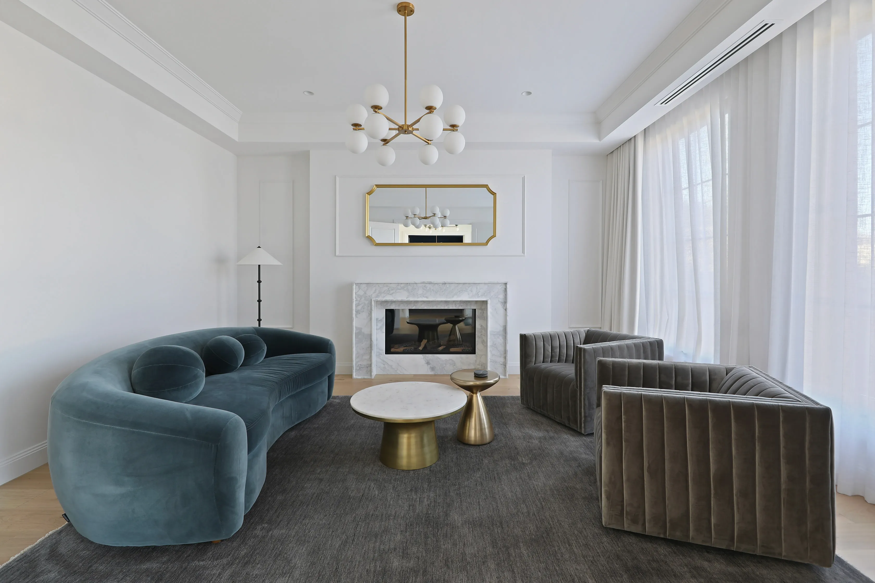 Formal living room with a white marble fireplace and gold-framed mirror, brass globe chandelier, a teal curved velvet sofa, fluted olive armchairs, brass-and-marble side tables on a grey rug