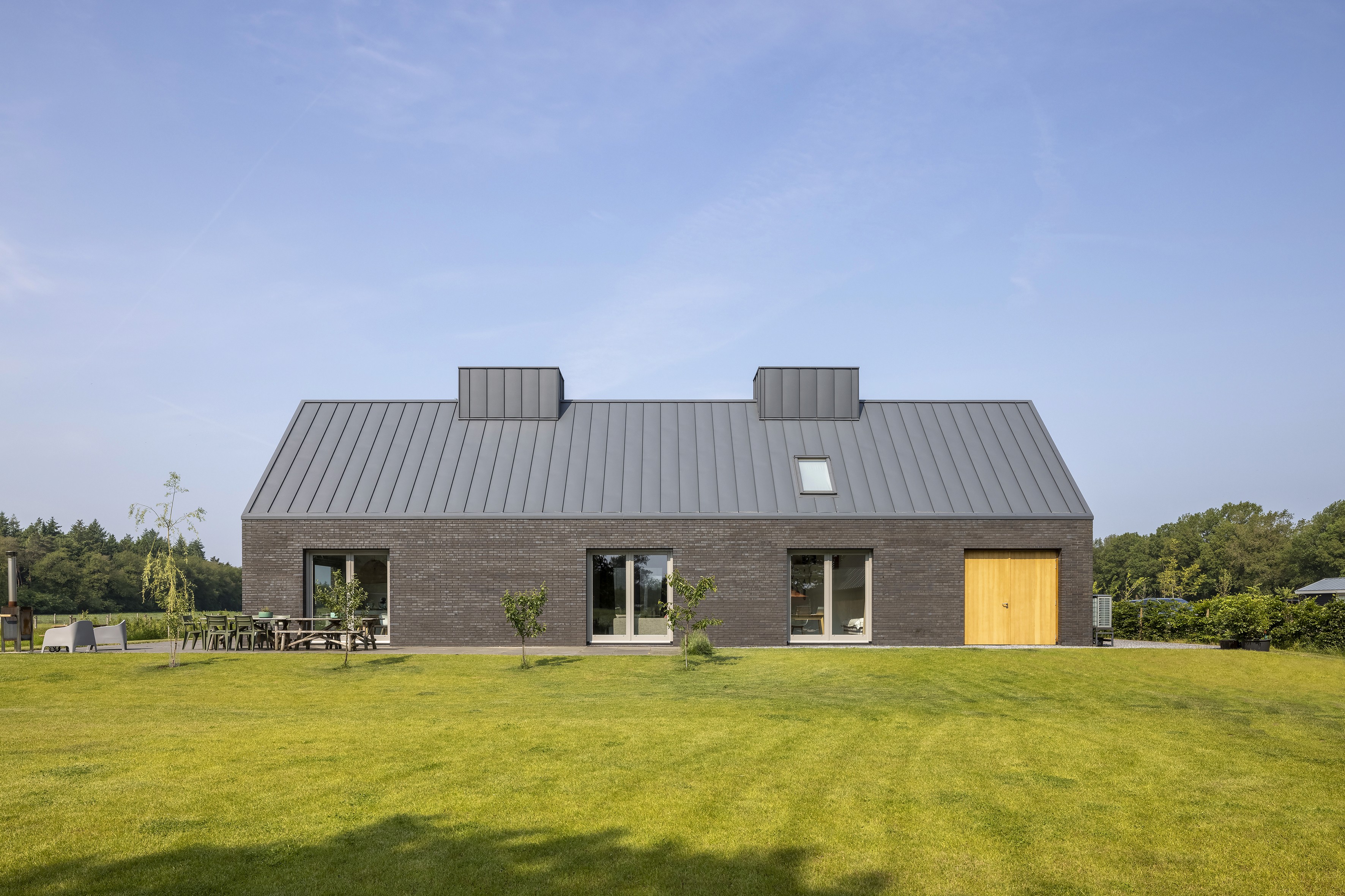 Modern huis met grijze baksteen, metalen dak en houten deur in groene omgeving.