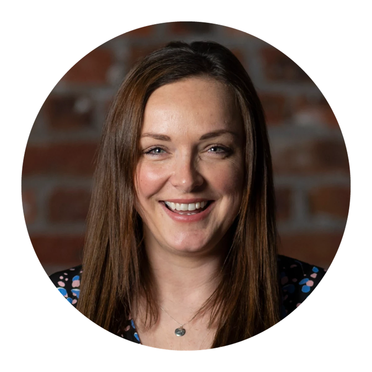 Person with straight brown hair wearing a dark top with colorful polka dots, standing in front of a brick wall background.