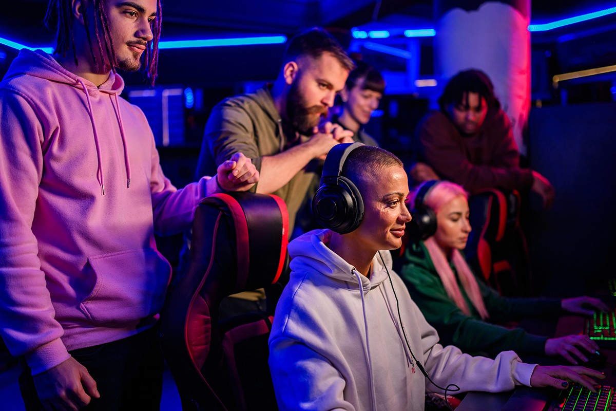 Group of people gathered around a row of computers in a dimly lit, neon-accented space. A person in a white hoodie focuses on the screen, while others stand nearby watching attentively