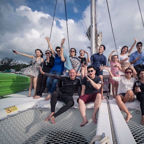 一群人在一个带网状座位区的帆船上微笑并充满活力地摆姿势，头顶是半云半晴的天空。