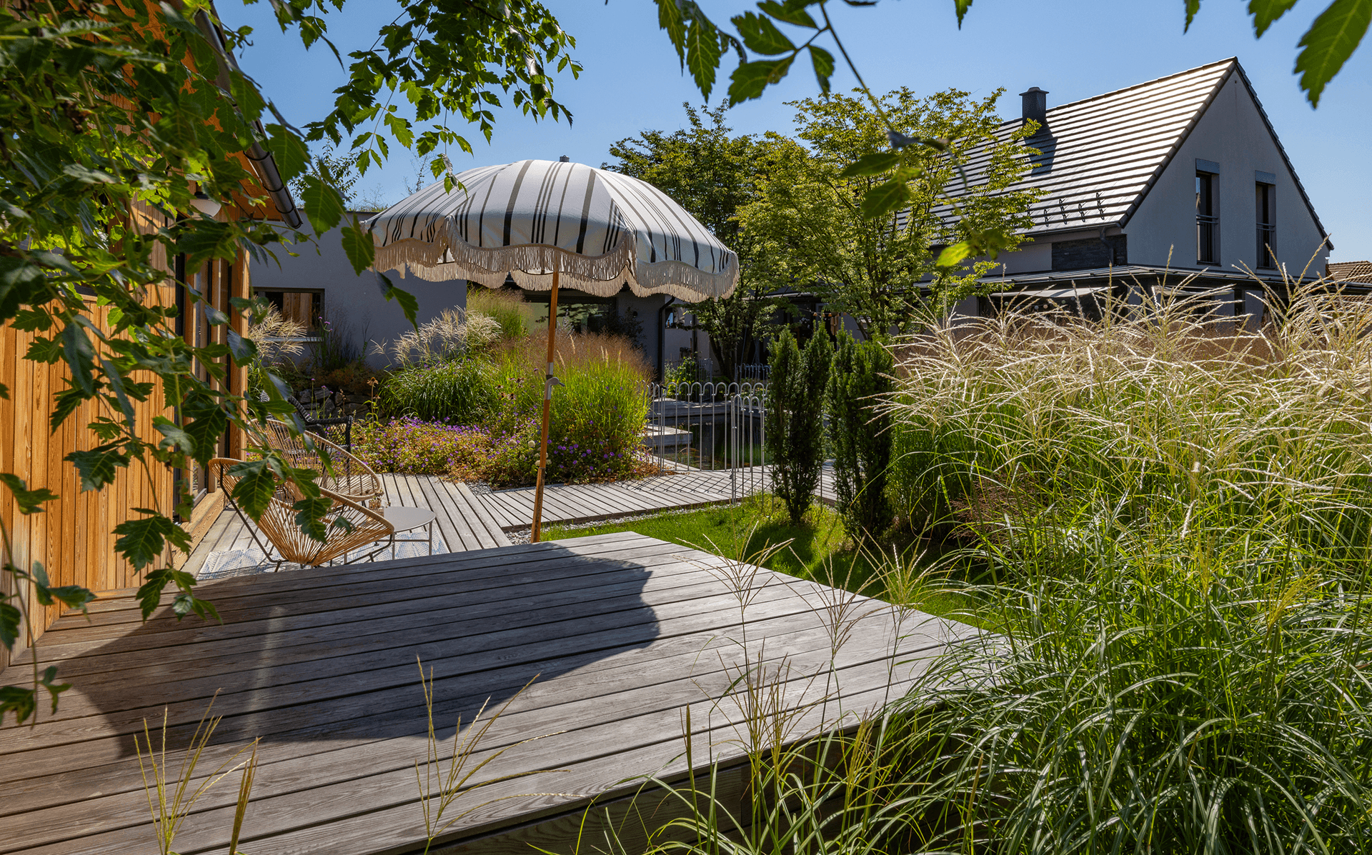 Holzterrasse mit Sonnenschirm neben einem natürlich gestalteten Teichgarten mit Gräsern und Stauden.