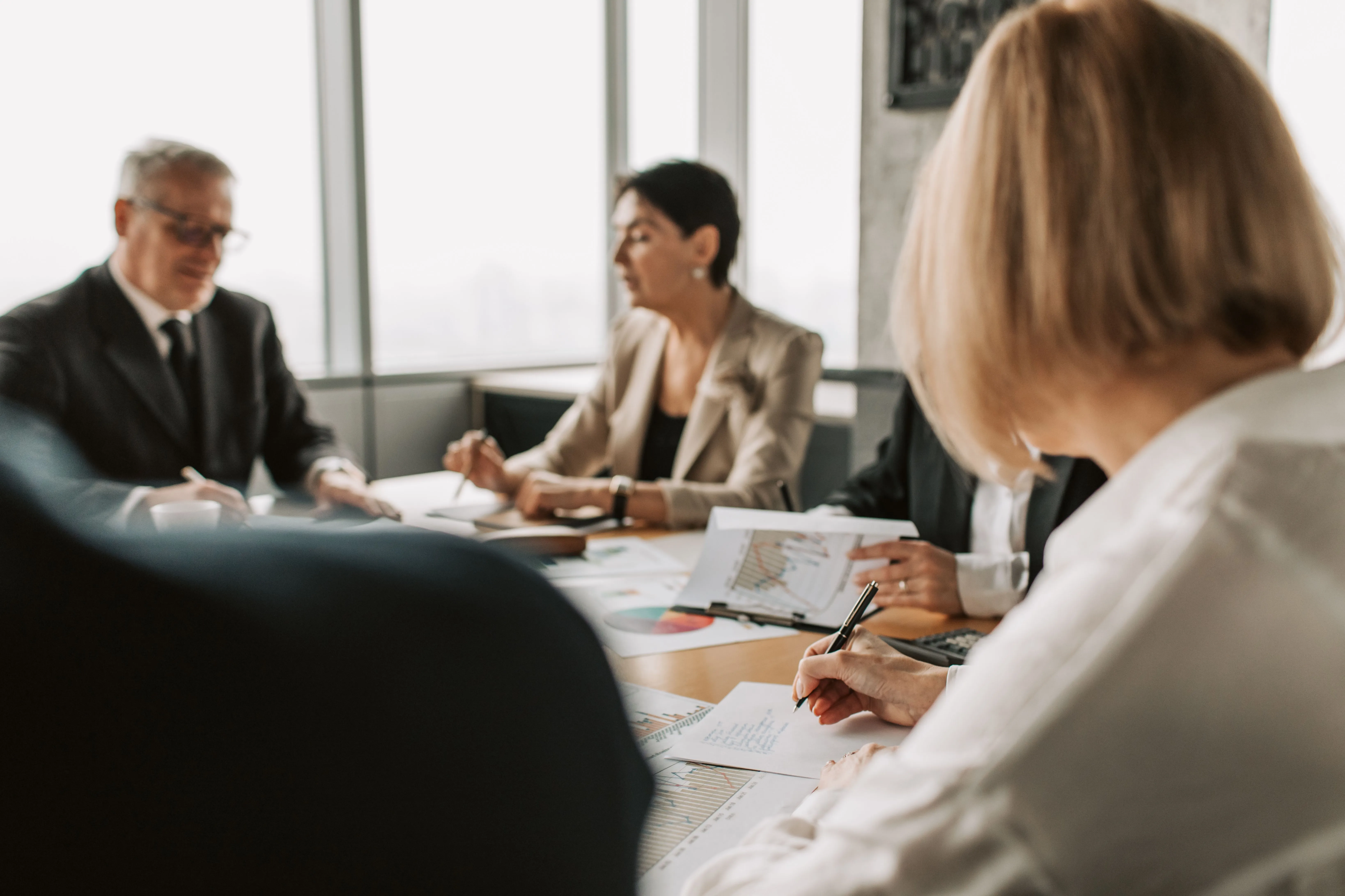 A group of professionals sits around a conference table in a bright office, reviewing printed charts and documents together, representing strategic business development and partnerships.