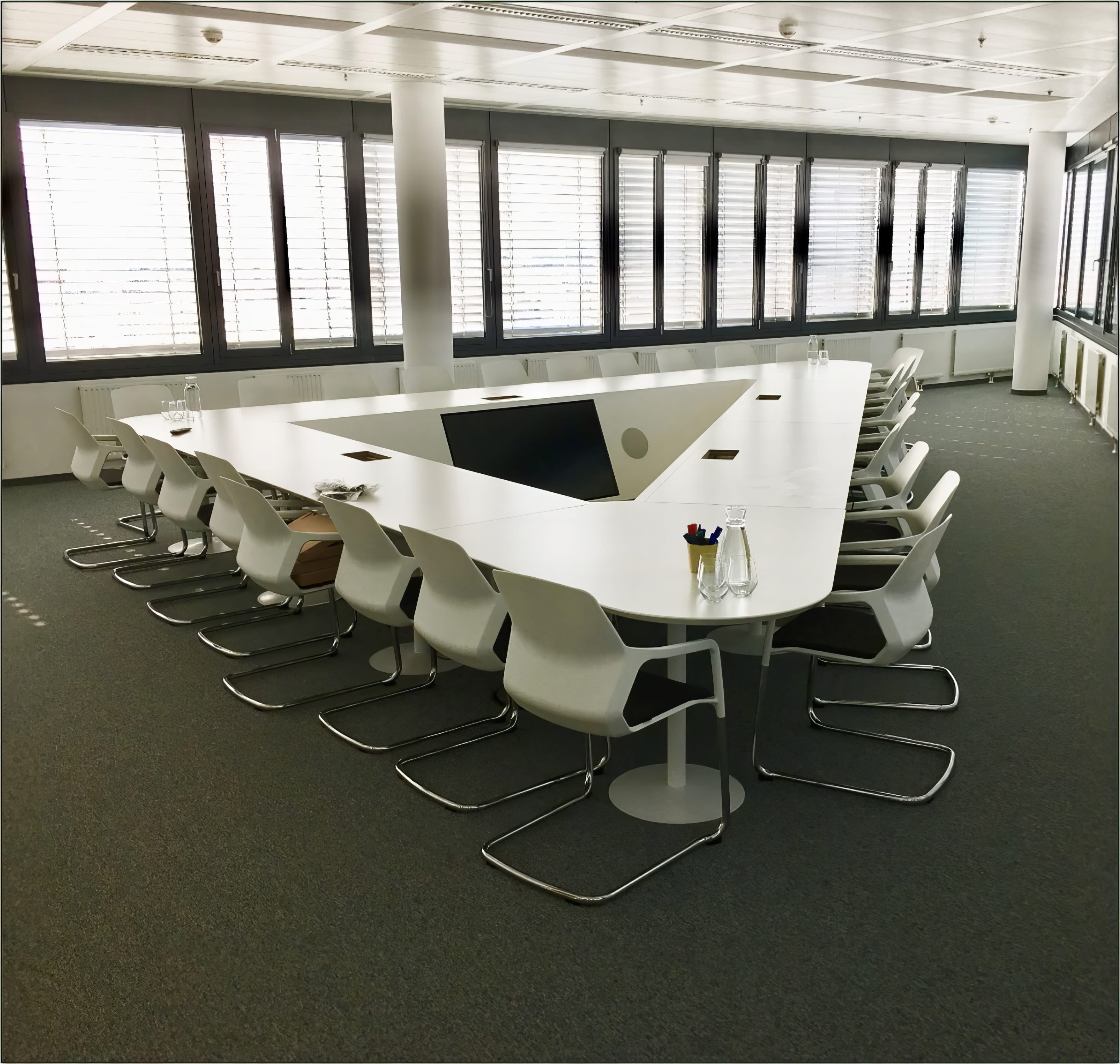MW Interior - Großer U-förmiger weißer Konferenztisch mit weißen Freischwingerstühlen, integriertem Display und umlaufender Fensterfront" Beschreibung: Großzügiger Konferenzraum mit maßgefertigtem U-Tisch in Weiß, integriertem versenkbarem Display und umlaufender Jalousien-Fensterfront – ideal für Präsentationen und Gruppenworkshops mit bis zu 20 Personen.