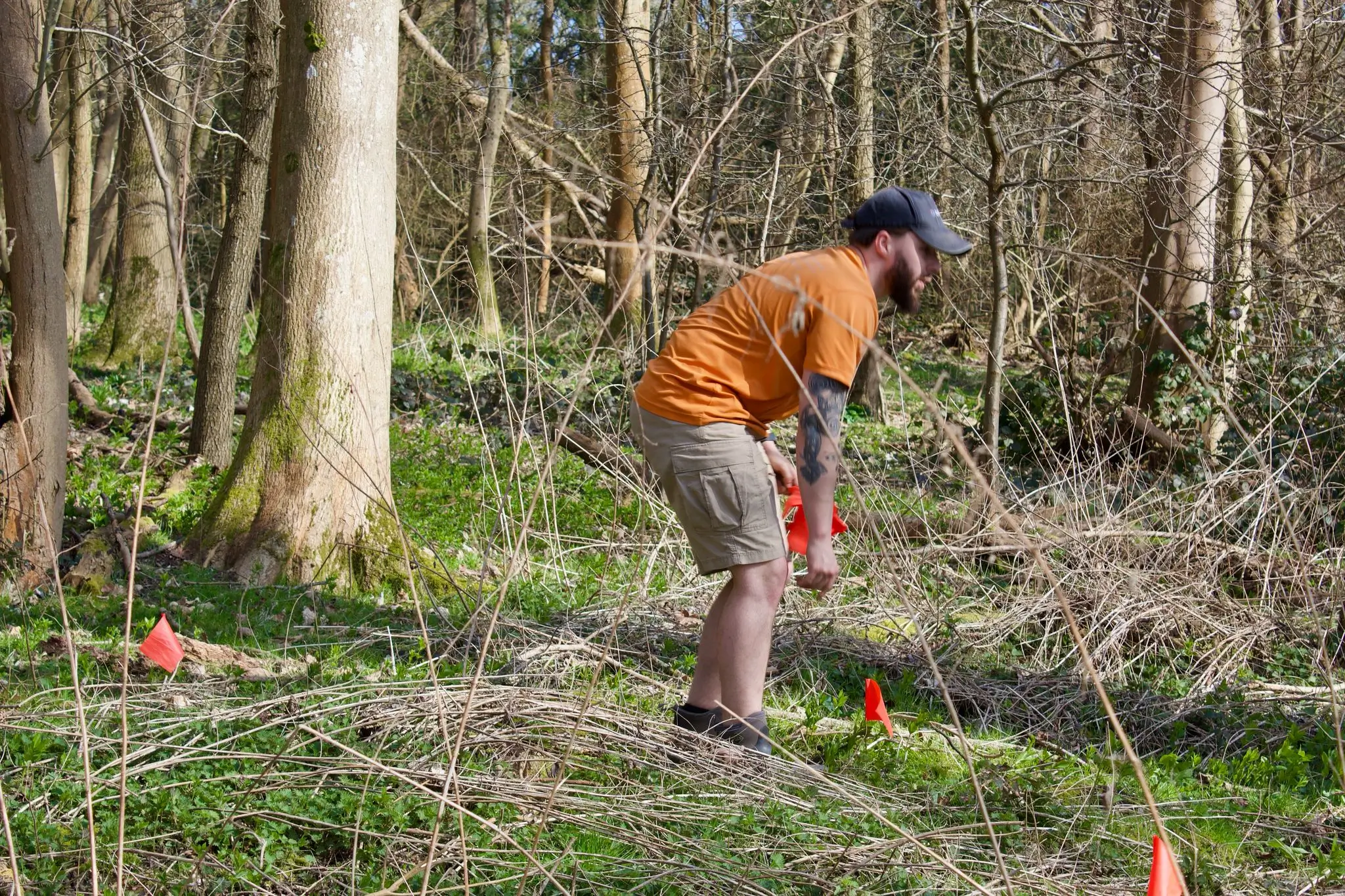 A person in an orange shirt bends down in a forested area, working on land or surveying with markers nearby.