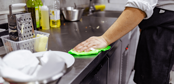 Person cleaning kitchen counter with cloth in commercial kitchen.
