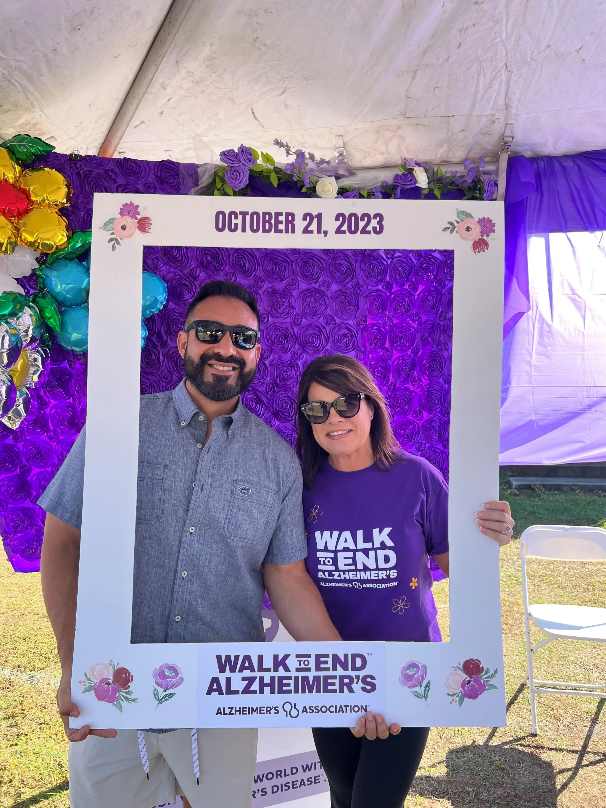 Two people wearing sunglasses pose together at a "Walk to End Alzheimer's" photo booth frame dated October 21, 2023, with purple rose decorations and colorful balloons in the background.