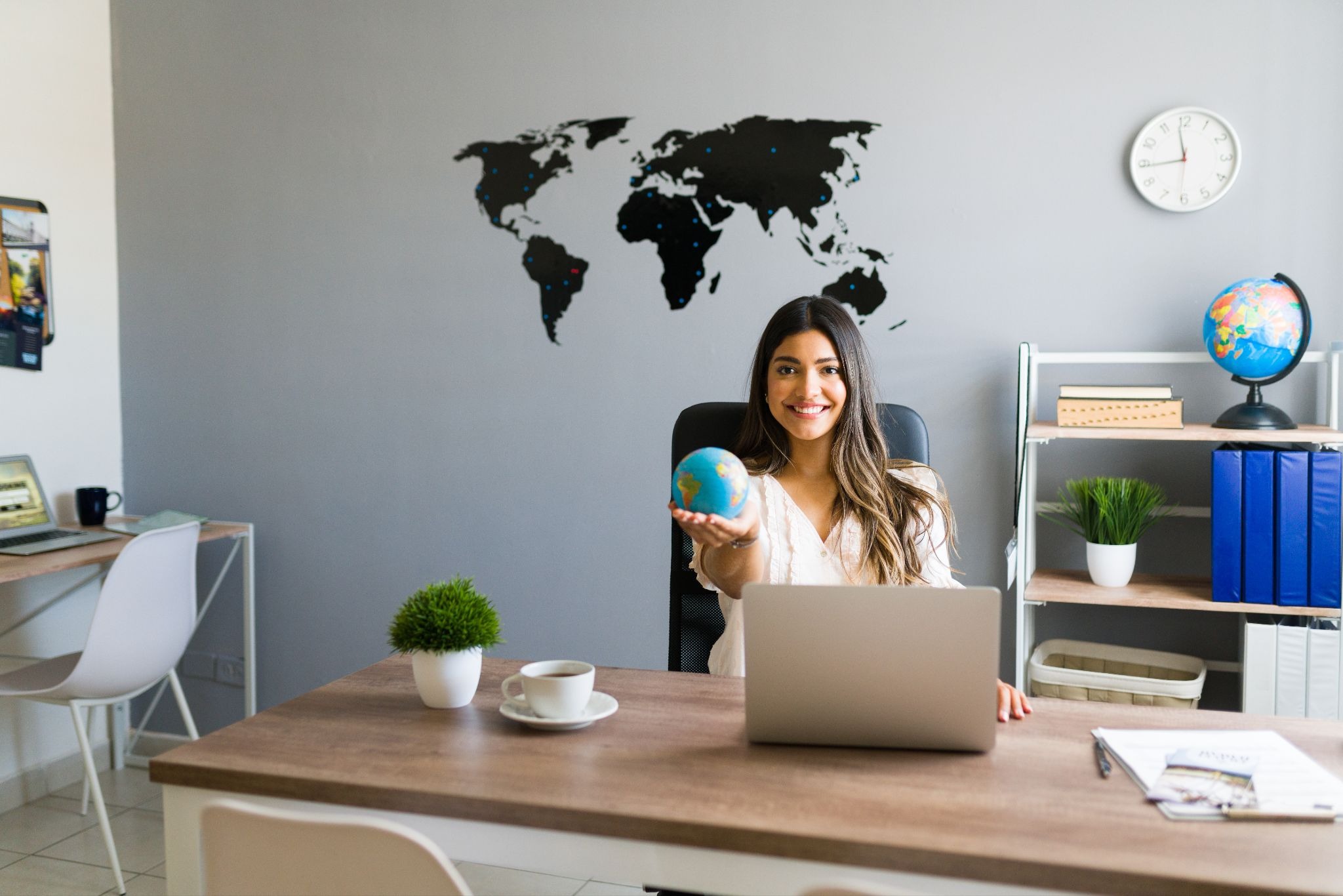 Smiling woman in an office holds a small globe, sitting at a desk with a laptop. World map and clock on wall, shelf with books and plant visible.