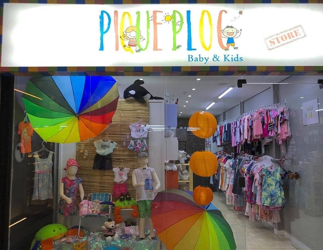 Fachada de loja infantil “Pique-Ploc Baby & Kids” com vitrine decorada por guarda-chuvas coloridos, manequins infantis e roupas expostas.