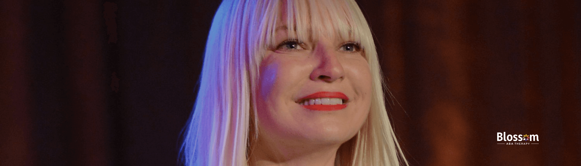 Sia smiled while revealing her autism diagnosis, wearing a blazer against a warm-lit background.