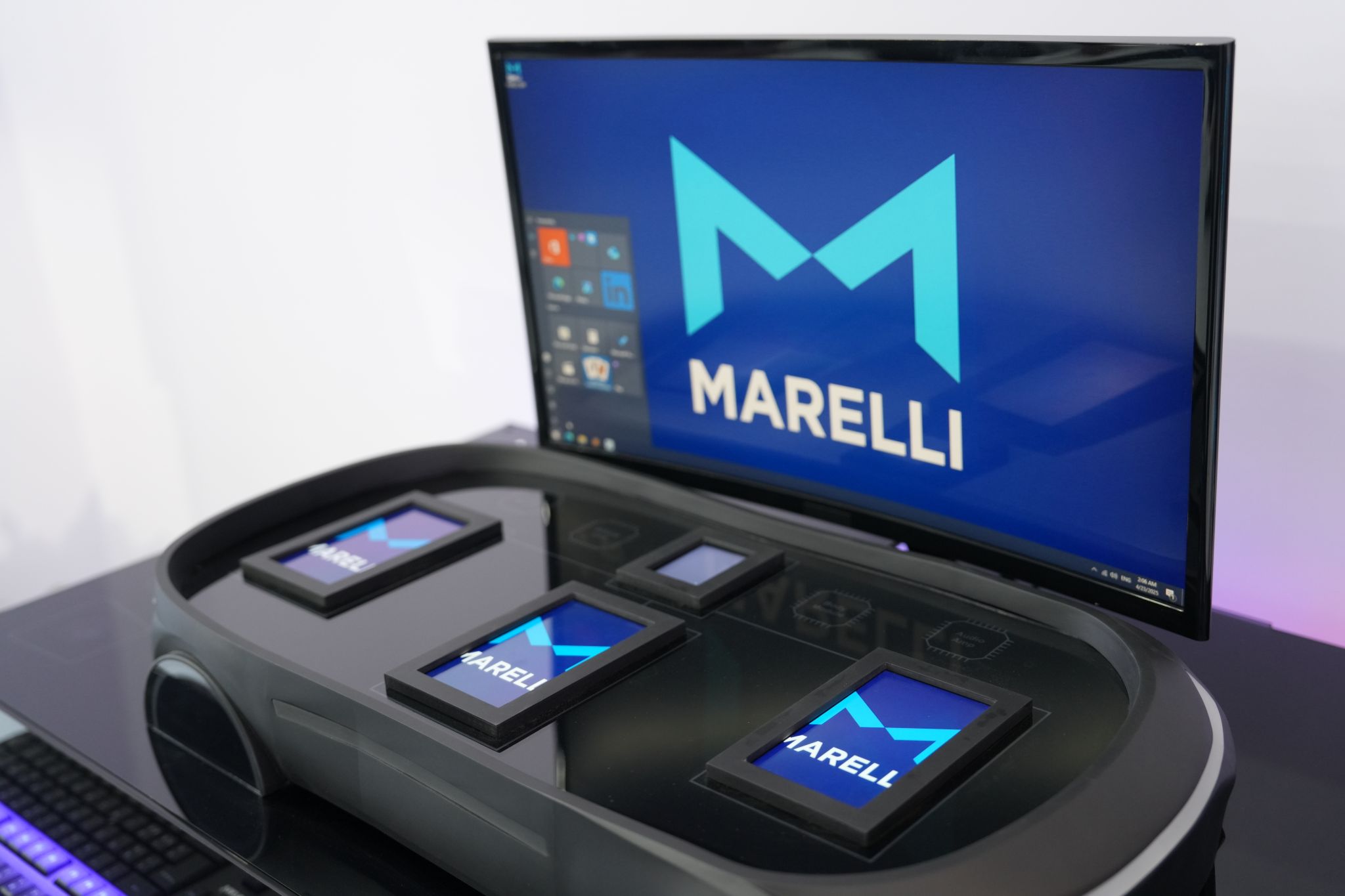 Postazione phygital Marelli: basamento auto in metallo satinato con dettagli cromati, tre monitor verticali su supporti sottili, palette scura con accenti arancioni, stile anni ’60–’70.