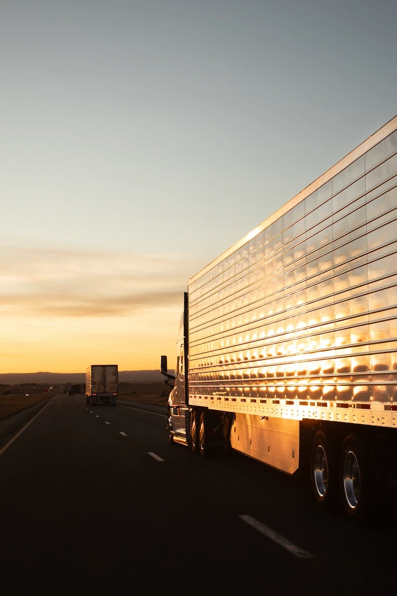 Refrigerated semi-truck trailer traveling on a highway at sunset, reflecting golden light off its polished aluminum sides.