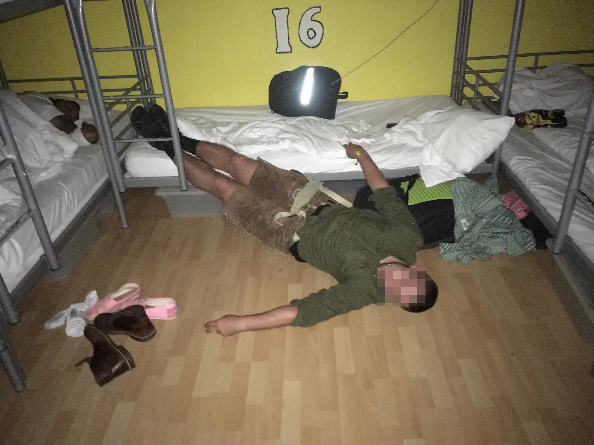 Man in lederhosen passed out on floor of hostel dorm with bunk beds, backpack, and shoes scattered.