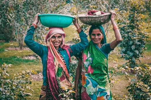 Two women carry baskets on their heads while walking through a lush, green landscape, wearing vibrant traditional clothing.