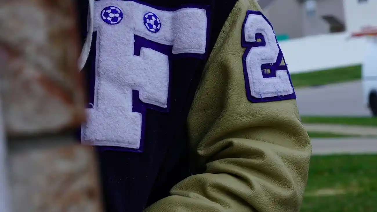 Close-up of a varsity-style jacket with football patches and stitched lettering, representing a sports club merch drop, sponsored apparel and community merchandise launch.