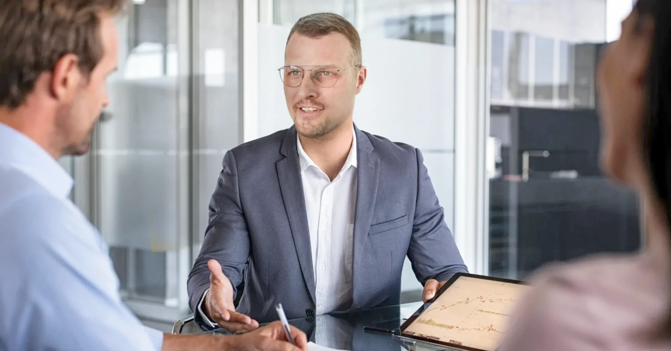 Geschäftsmann im Anzug erklärt etwas während eines Meetings und zeigt dabei auf ein Tablet mit Diagrammen, zwei Kollegen hören ihm zu.