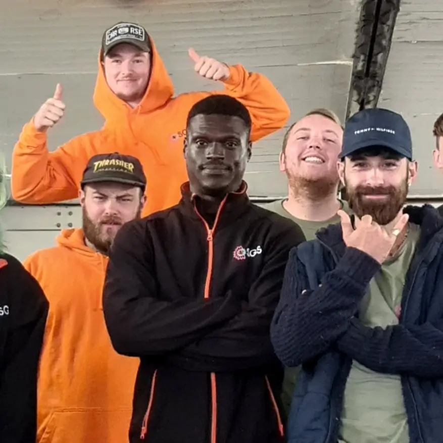 A group of men posing together, some smiling, wearing various hats and jackets, with an industrial background.