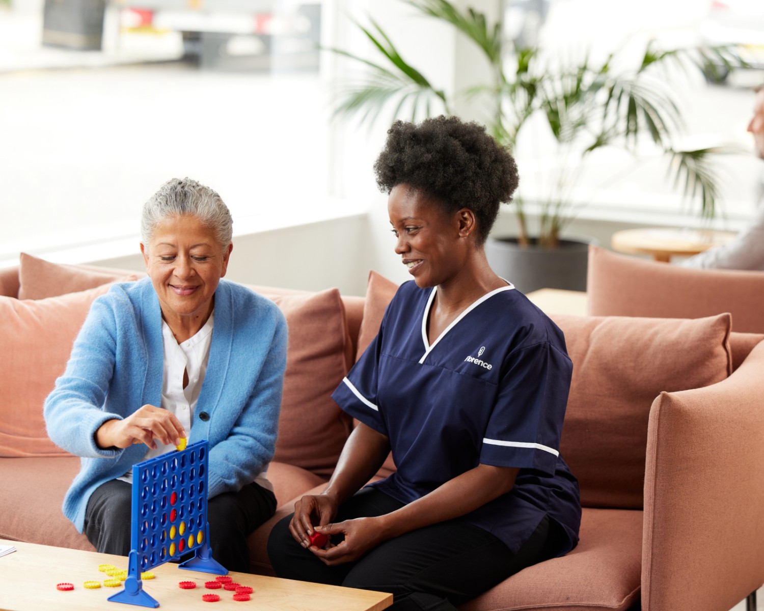 Older woman and a Florence caregiver sitting on a sofa playing a board game together in a bright, comfortable living space.