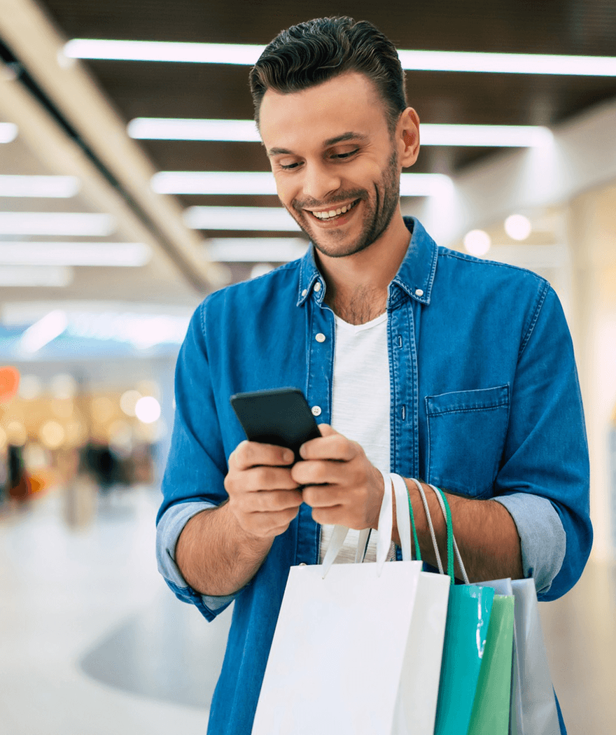 A man holding shopping bags while checking his phone, appearing engaged and focused on his device.