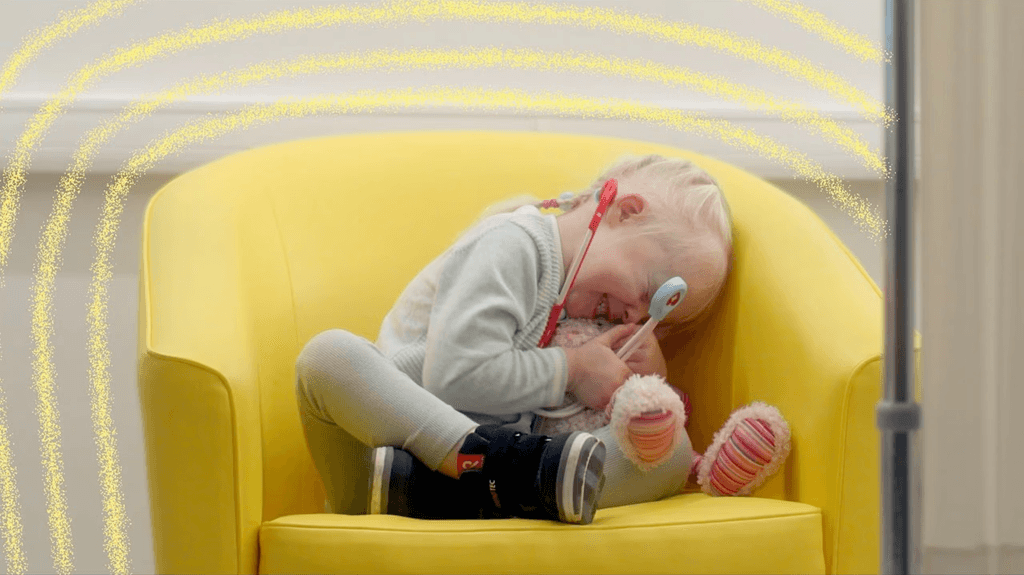 Screenshot from the ad, showing a small girl hugging a stuffed bunny on a chair with a toy stethoscope in her hand, and some sparkly lines eminating from her (my animations).